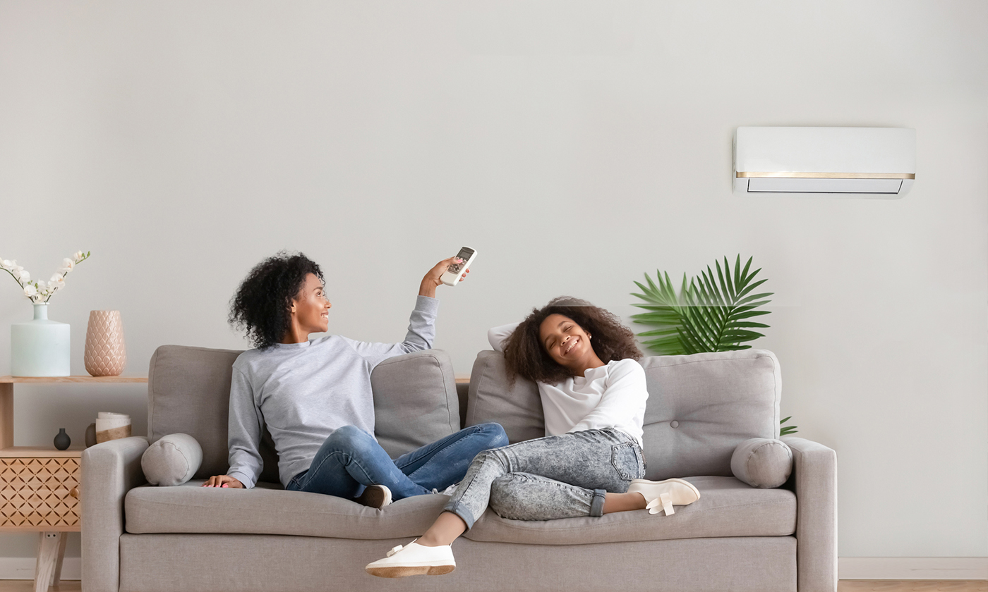 A mum and daughter sitting on a couch smiling, mum is using a remote control to adjust the split system